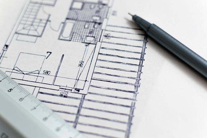 A close up of an architects blue prints with a pen and ruler slightly out of focus ontop of the blueprints