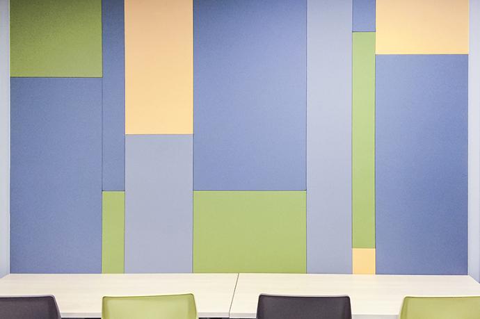 A Decorative Fabricmate Fabric Wall installation with a desk and chairs in the foreground. The Fabricmate wall features vertical bands with various widths and heights. The color of the fabric wall sections is yellow, dark blue, light blue and green.