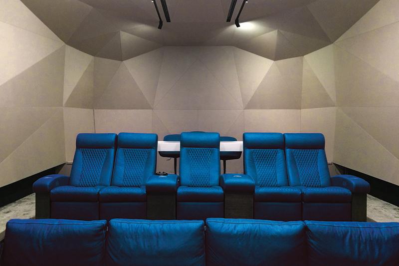 A dramatically lit home theater system with Fabricmate wall installations. The home theater has 2 rows of blue comfortable seats and the walls have geometric shaped wall installations that are beige color and are lit with floodlights.