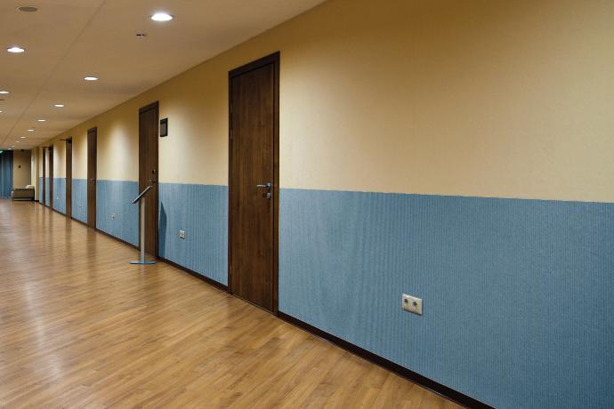 A hallway with many closed brown doors. The walls feature Fabricmate wall installations. The bottom half of the fabric wall is light blue and the top is a cream color.